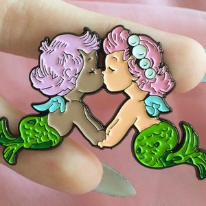 May include: A pin depicting two mermaids in an embrace. One mermaid has pink hair, the other has brown skin and pink hair. Both have green tails and blue accents. The pin is black with colourful enamel.
