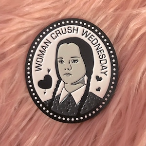 May include: A round, black and white pin with the text "WOMAN CRUSH WEDNESDAY" encircling a portrait of Wednesday Addams. The pin features a black heart pierced by an arrow and two small hearts.