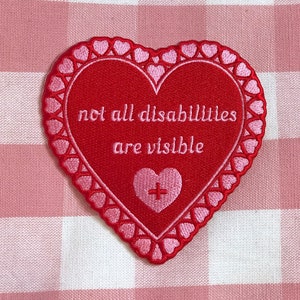 Könnte beinhalten: Gestickter herzförmiger Aufnäher in Rot mit dem Text „not all disabilities are visible“. Der Aufnäher hat einen rosa Herzrand und ein kleineres rosa Herz mit einem Pluszeichen. Auf rosa-weiß kariertem Hintergrund.