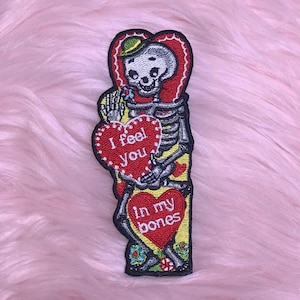 May include: Embroidered patch featuring a skeleton holding two red heart-shaped cards. The top card reads "I feel you," and the bottom card says "In my bones." The skeleton wears a green hat and is surrounded by floral details.