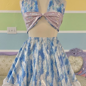 Puede incluir: Un conjunto de dos piezas con top halter y falda. El top es azul y blanco con un lazo. La falda es azul y blanca con un ribete de encaje inglés blanco. El conjunto se exhibe en un maniquí.