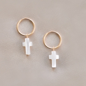May include: A pair of gold hoop earrings with a white cross charm hanging from each hoop.