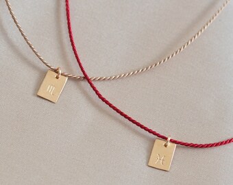May include: Two gold zodiac symbol pendants on red and beige cords. The pendant on the red cord is a Pisces symbol, and the pendant on the beige cord is a Gemini symbol.