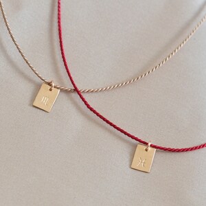 May include: Two gold zodiac symbol pendants on red and beige cords. The pendant on the red cord is a Pisces symbol, and the pendant on the beige cord is a Gemini symbol.