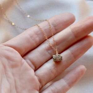 Puede incluir: Un delicado collar de oro con un pequeño colgante de pirita texturizada. El colgante tiene forma de cubo con un aspecto metálico y moteado. El collar se muestra en una mano sobre un fondo suave y neutro.