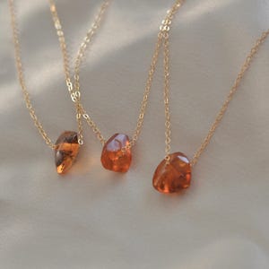 May include: Three gold necklaces, each featuring a unique, irregularly shaped amber pendant. The amber stones vary in size and hue, ranging from light orange to a deeper, reddish-brown. The necklaces are displayed on a soft, off-white fabric.