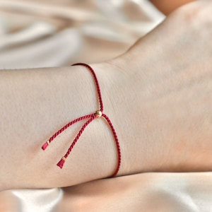 Puede incluir: Un brazalete de cuerda roja con un acento de cuentas doradas. El brazalete está atado con un nudo y tiene dos extremos sueltos.