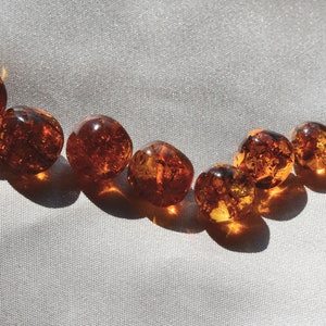 Peut inclure: Une collection de perles d'ambre de diff&eacute;rentes nuances de brun et d'orange, dispos&eacute;es en ligne courbe sur un fond de tissu blanc.