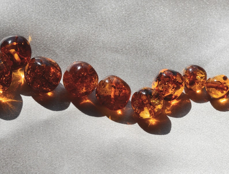 Peut inclure: Une collection de perles d'ambre de diff&eacute;rentes nuances de brun et d'orange, dispos&eacute;es en ligne courbe sur un fond de tissu blanc.