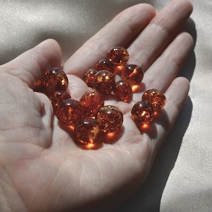 Peut inclure: Une main tenant une collection de perles d'ambre. Les perles sont rondes et translucides, d'une couleur chaude et miell&eacute;e. Elles sont dispos&eacute;es en un tas l&acirc;che, avec quelques chevauchements.