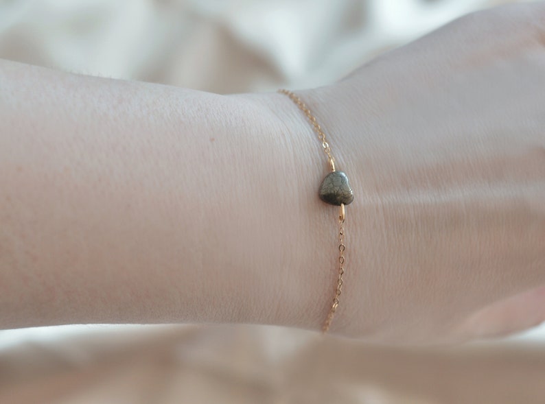 Puede incluir: Una delicada pulsera de cadena dorada con una peque&ntilde;a piedra en forma de coraz&oacute;n, de color gris verdoso. La pulsera es sencilla y elegante, con un dise&ntilde;o minimalista. La piedra es el punto focal, a&ntilde;adiendo un toque de belleza natural a la joya.