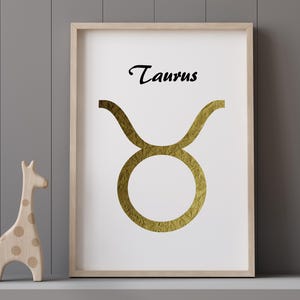 May include: A framed print featuring the Taurus zodiac symbol in gold foil on a white background. The word "Taurus" is written in black script above the symbol. A wooden giraffe figurine stands to the left of the framed print.