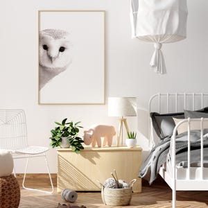 May include: A white owl print in a wooden frame hangs on a white wall above a wooden side table. The table has a potted plant, a pink toy animal, and a white lamp on it. A white metal bed frame with a gray and white striped blanket is in the background.