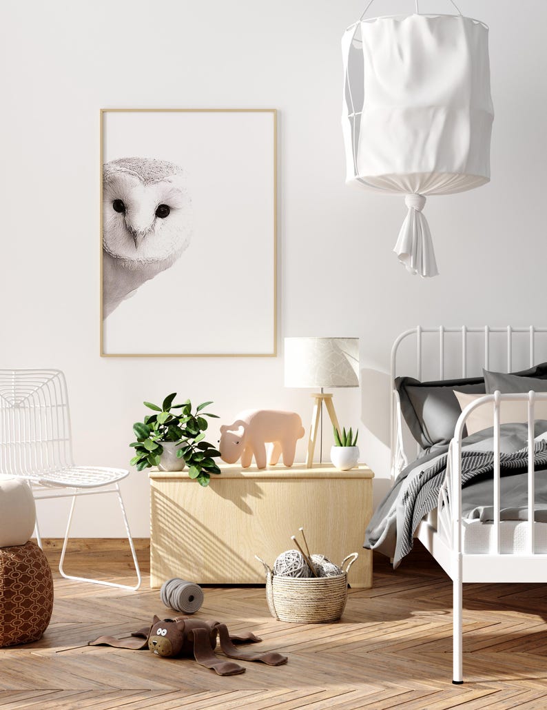 May include: A white owl print in a wooden frame hangs on a white wall above a wooden side table. The table has a potted plant, a pink toy animal, and a white lamp on it. A white metal bed frame with a gray and white striped blanket is in the background.