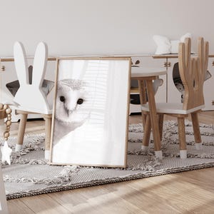May include: A white bunny-shaped chair and a wooden deer-shaped chair with white seats are arranged around a small wooden table. A framed print of a white owl with black eyes is leaning against the table. The chairs and table are on a light gray rug.