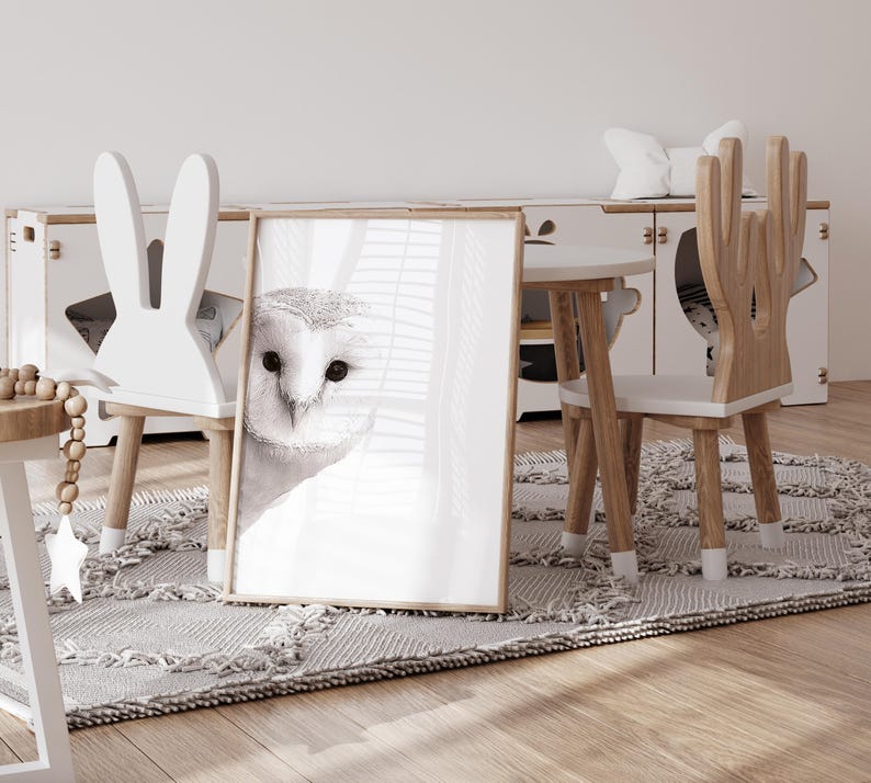 May include: A white bunny-shaped chair and a wooden deer-shaped chair with white seats are arranged around a small wooden table. A framed print of a white owl with black eyes is leaning against the table. The chairs and table are on a light gray rug.