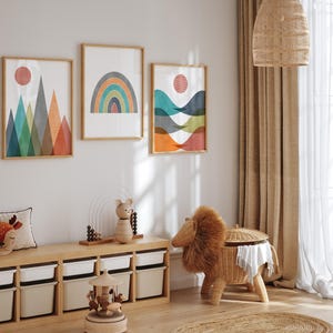 May include: Three abstract geometric wall art prints featuring a rainbow, mountains, and waves. The prints are framed in natural wood and have a white background. The prints are hanging on a white wall above a wooden shelf with storage bins.