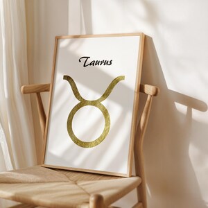 May include: Framed art print featuring the Taurus zodiac symbol in gold against a white background. The word "Taurus" is written in black script above the symbol. The artwork is displayed on a wooden chair.