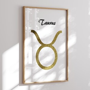 May include: Framed art print featuring the Taurus zodiac symbol. The gold-colored symbol is set against a white background, with the word "Taurus" in black script above. The artwork is encased in a light-colored wooden frame, suitable for home decor.