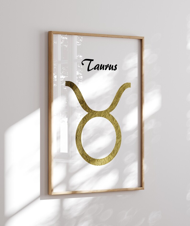 May include: Framed art print featuring the Taurus zodiac symbol. The gold-colored symbol is set against a white background, with the word "Taurus" in black script above. The artwork is encased in a light-colored wooden frame, suitable for home decor.