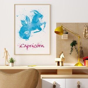 May include: A framed watercolor print featuring the Capricorn zodiac sign. The artwork depicts a blue mythical creature with a fish tail and goat horns, with the word "Capricorn" in pink script below. The print is displayed on a desk.