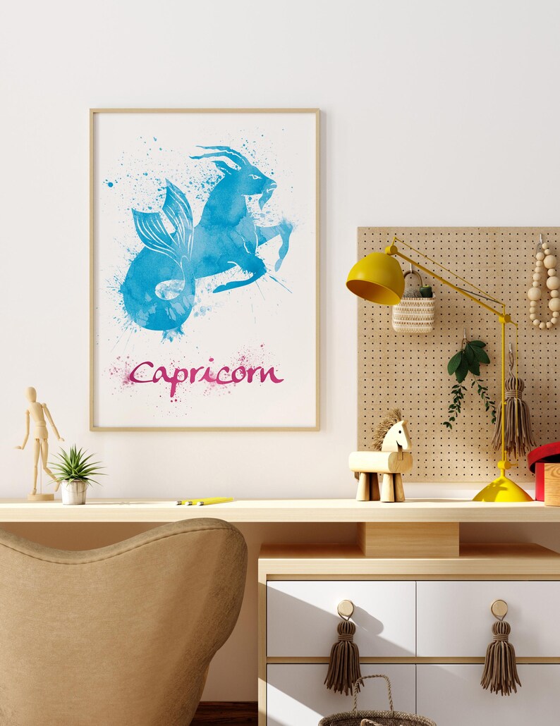 May include: A framed watercolor print featuring the Capricorn zodiac sign. The artwork depicts a blue mythical creature with a fish tail and goat horns, with the word "Capricorn" in pink script below. The print is displayed on a desk.