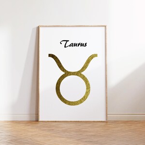 May include: A framed print featuring the Taurus zodiac symbol in gold against a white background. The word "Taurus" is written in black script above the symbol. The artwork is displayed on a wooden floor.