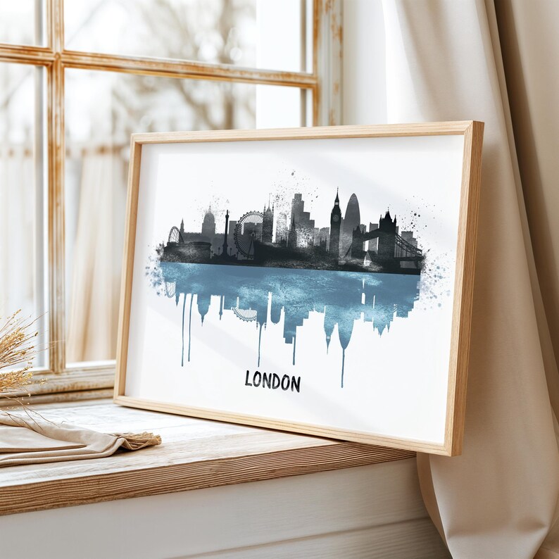 May include: A framed art print featuring a watercolor silhouette of the London skyline in shades of gray and blue. The word "LONDON" is printed in black at the bottom. The print is displayed on a wooden windowsill.