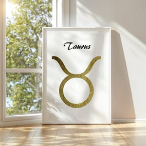 May include: A framed white print featuring the word "Taurus" in elegant black script above a gold-colored zodiac symbol. The artwork is displayed against a white wall, with natural light illuminating the scene.