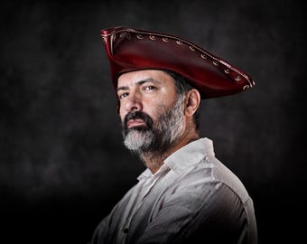 May include: A deep red, structured tricorn hat featuring visible stitching along the brim edges. The hat has a curved, triangular shape with a smooth, polished finish, designed to be worn on the head with a distinct, historical aesthetic.