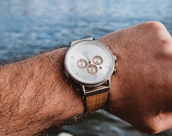 May include: A silver chronograph wristwatch with a brown leather band. The watch is on a person's wrist, with the background out of focus.