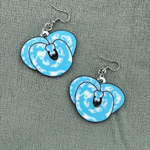 Peut inclure: Une paire de boucles d'oreilles représentant un motif de serpent bleu et blanc. Les boucles d'oreilles sont en forme de cœur avec un contour noir et des crochets argentés. Le motif du serpent est bleu clair avec des taches blanches.