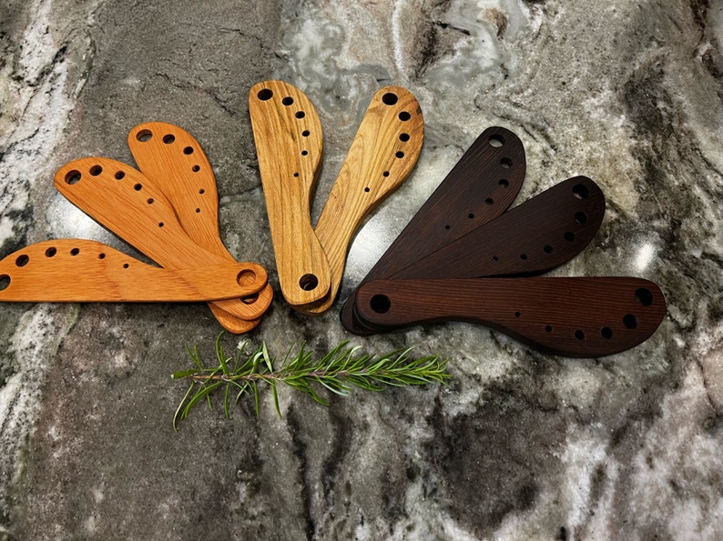 This image includes eight handmade wooden herb strippers, three cherry, two teak and three walnut. These herb strippers include incrementally larger holes that a cook can use to pull herbs through to separate the stems from the leaves.