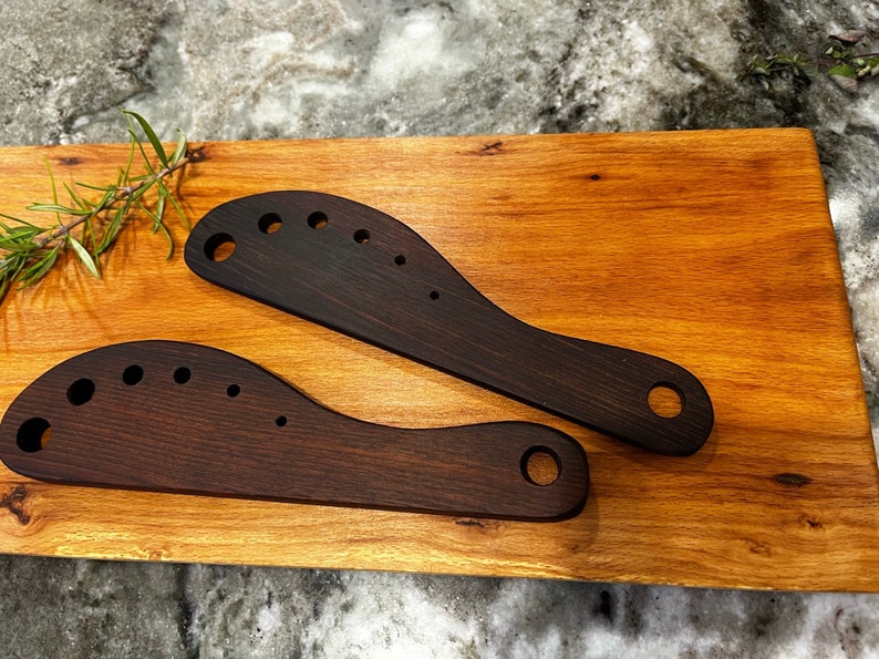 This image includes two handmade herb strippers made from walnut. 

These herb strippers include incrementally larger holes that a cook can use to pull herbs through to separate the stems from the leaves.
