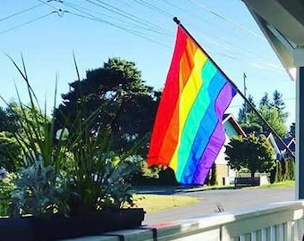 May include: A rainbow flag, representing the LGBTQ+ community, is displayed on a porch. The flag is attached to a black pole and hangs in front of a blue sky. The porch has white railings and a small wooden table.