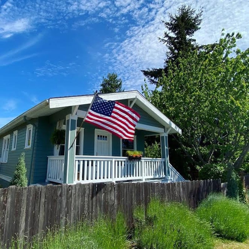 May include: A blue house with a white porch and a white picket fence. An American flag is flying in front of the house. The house is surrounded by green grass and trees.