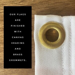 May include: Our flags are finished with canvas heading and brass grommets. A close-up of a white fabric flag with a brass grommet.