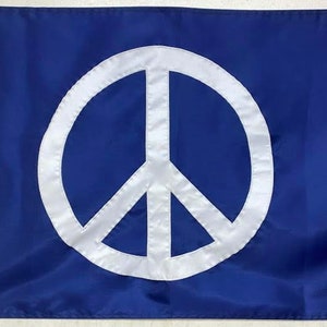 Puede incluir: Bandera azul con un símbolo de la paz blanco en el centro. El símbolo de la paz es un círculo con tres líneas hacia abajo. La bandera tiene un borde blanco en el borde izquierdo y es de material liso. Símbolo de la paz.