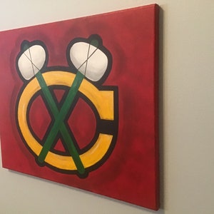May include: A red, yellow, green, and white canvas painting of the Chicago Blackhawks logo. The logo features a stylized "C" with two crossed hockey sticks and a white puck on each stick.