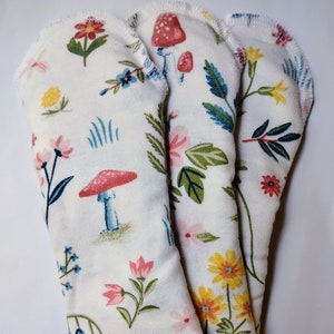 May include: Three reusable cloth menstrual pads with a white background and a floral print featuring mushrooms, flowers, and leaves in shades of pink, yellow, blue, and green.