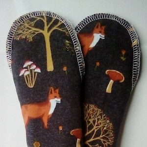 以下が含まれることがあります： 白のジグザグステッチが縁取りされた、濃いグレーの布製の鍋つかみが2つ。布地には、オレンジ色のキツネ、黄色の木、茶色のキノコが繰り返し描かれた模様があります。