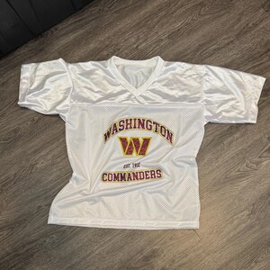 May include: White mesh football jersey with a V-neck and short sleeves. The jersey features the text "WASHINGTON COMMANDERS" in gold and burgundy, with a burgundy "W" logo. The text "EST. 1932" is also visible.