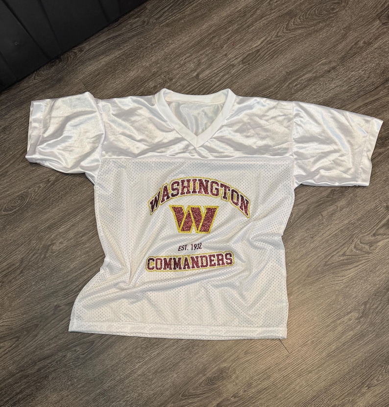 May include: White mesh football jersey with a V-neck and short sleeves. The jersey features the text "WASHINGTON COMMANDERS" in gold and burgundy, with a burgundy "W" logo. The text "EST. 1932" is also visible.