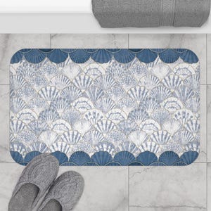 May include: A rectangular bath mat with a seashell pattern in shades of blue and white. The mat has a scalloped border in a darker blue. Gray slippers are at the bottom of the mat. A gray towel is at the top.