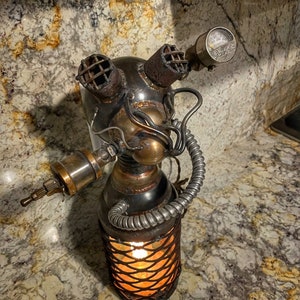 May include: A steampunk-style lamp with a metal cage base and a bulb inside, emitting a warm glow. The lamp features a diver's helmet design with various metal components, including gauges and vents. The lamp is made of copper, brass, and steel.