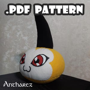 Puede incluir: Un peluche con forma de personaje de dibujos animados con cara blanca, cuerpo amarillo y cuerno negro. El personaje tiene grandes ojos rojos y una boca sonriente. El texto ".PDF PATTERN" está en la parte superior y "Antharez" en la inferior.
