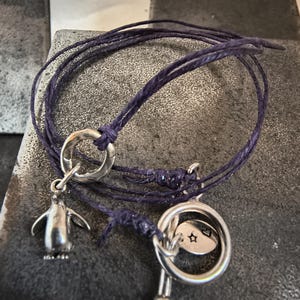 May include: A dark purple cord bracelet with a silver penguin charm and a silver toggle clasp. The bracelet is knotted and has a small silver charm with a star. The bracelet is on a textured surface.