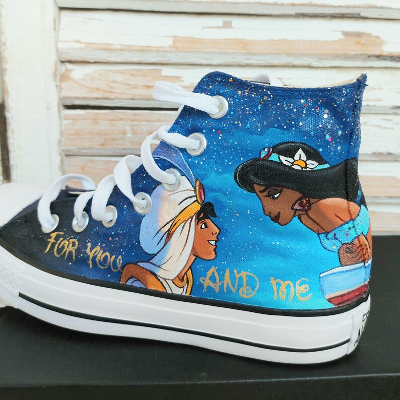 Op de afbeelding: Op maat geschilderde hoge canvas sneakers met een sc&egrave;ne uit Aladdin. Het ontwerp bevat Aladdin en prinses Jasmine tegen een sterrenblauwe achtergrond, met de tekst "FOR YOU AND ME" in gouden letters. De schoenen hebben witte veters en een zwarte zool.