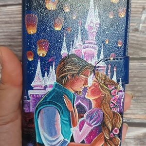 Puede incluir: Una funda de teléfono con un diseño pintado a mano de una pareja besándose frente a un castillo. El castillo está rodeado de linternas flotantes y el cielo es azul oscuro.