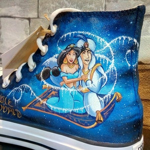 Op de afbeelding: Hoge canvas schoen met een handgeschilderd ontwerp van Aladdin en Jasmine op een vliegend tapijt tegen een sterrenblauwe achtergrond. De schoen heeft witte veters en een witte zool. De woorden "WHOLE WORLD" zijn zichtbaar.